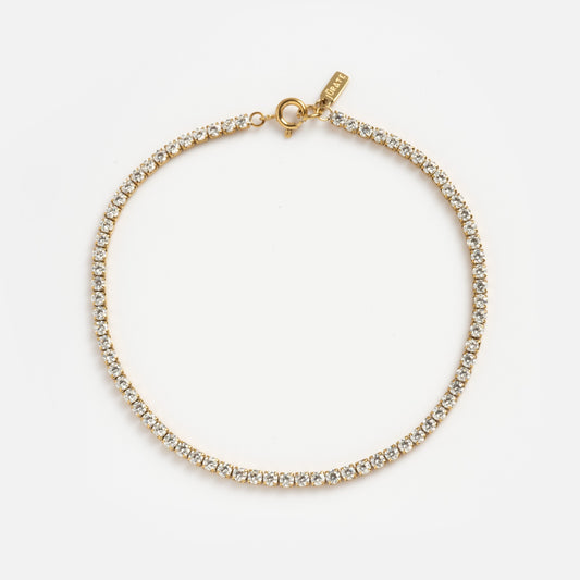 A gold tennis necklace with clear, round-cut gemstones and a lobster clasp is shown on a white background—pairs perfectly with the Material Girl Anklet for an extra hint of luxury.