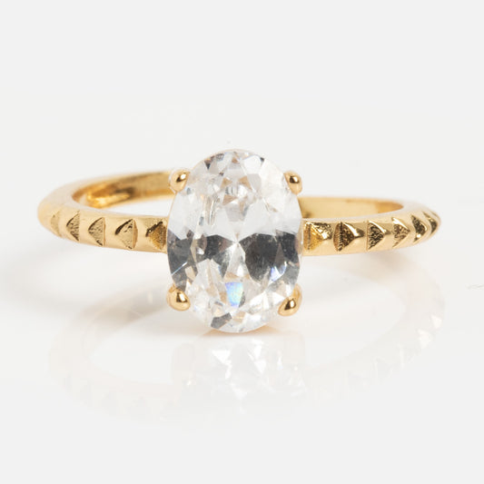 The Still Toe Ring is a luxury 18k gold plated piece featuring a large oval clear gemstone in four prongs and a textured band with small triangular engravings for an elegant finish.