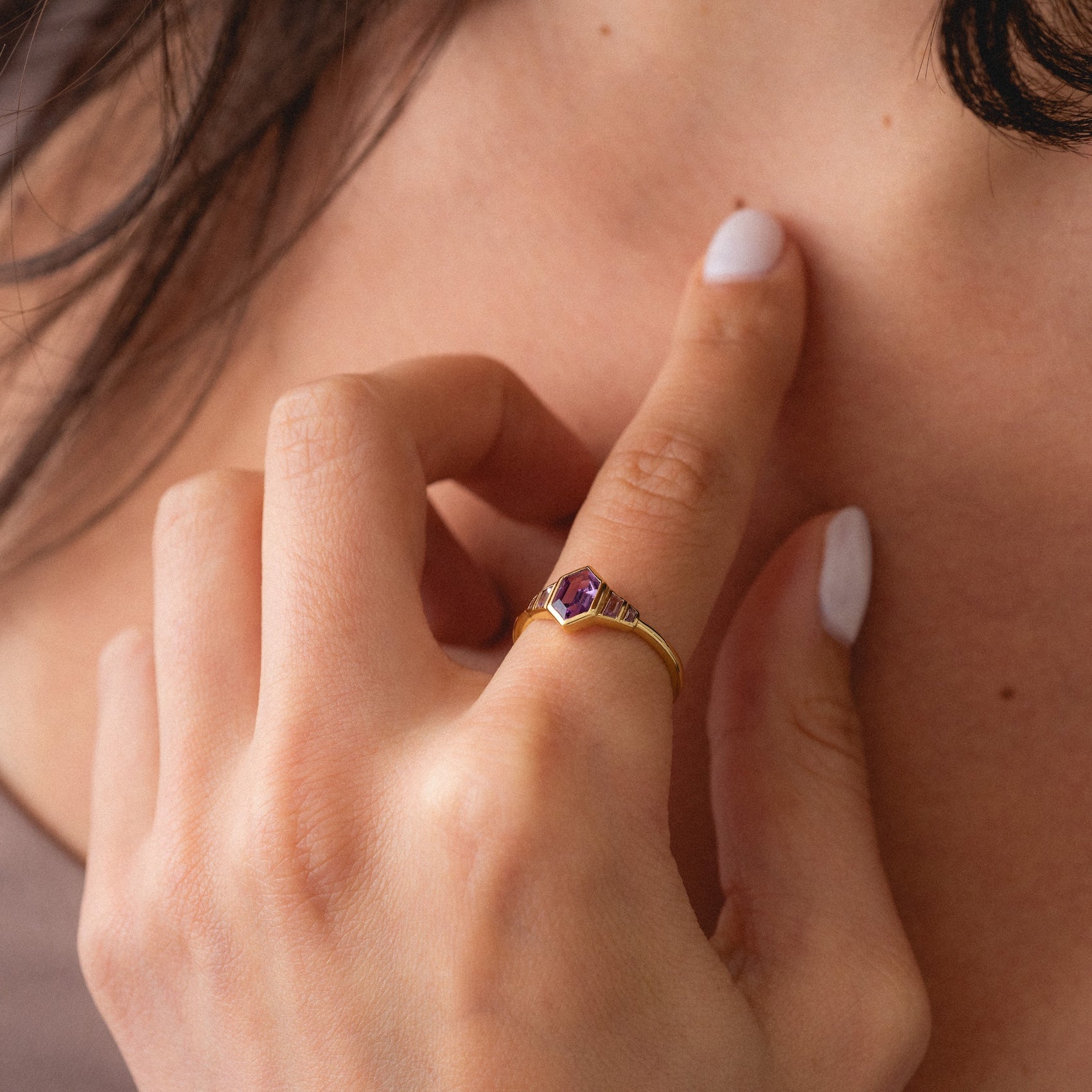 A close-up of manicured fingers touching a collarbone highlights the 2026 June Capsule Art Deco Alexandrite Ring, featuring a gold setting with a purple gemstone. Strands of dark hair frame the top of the image.