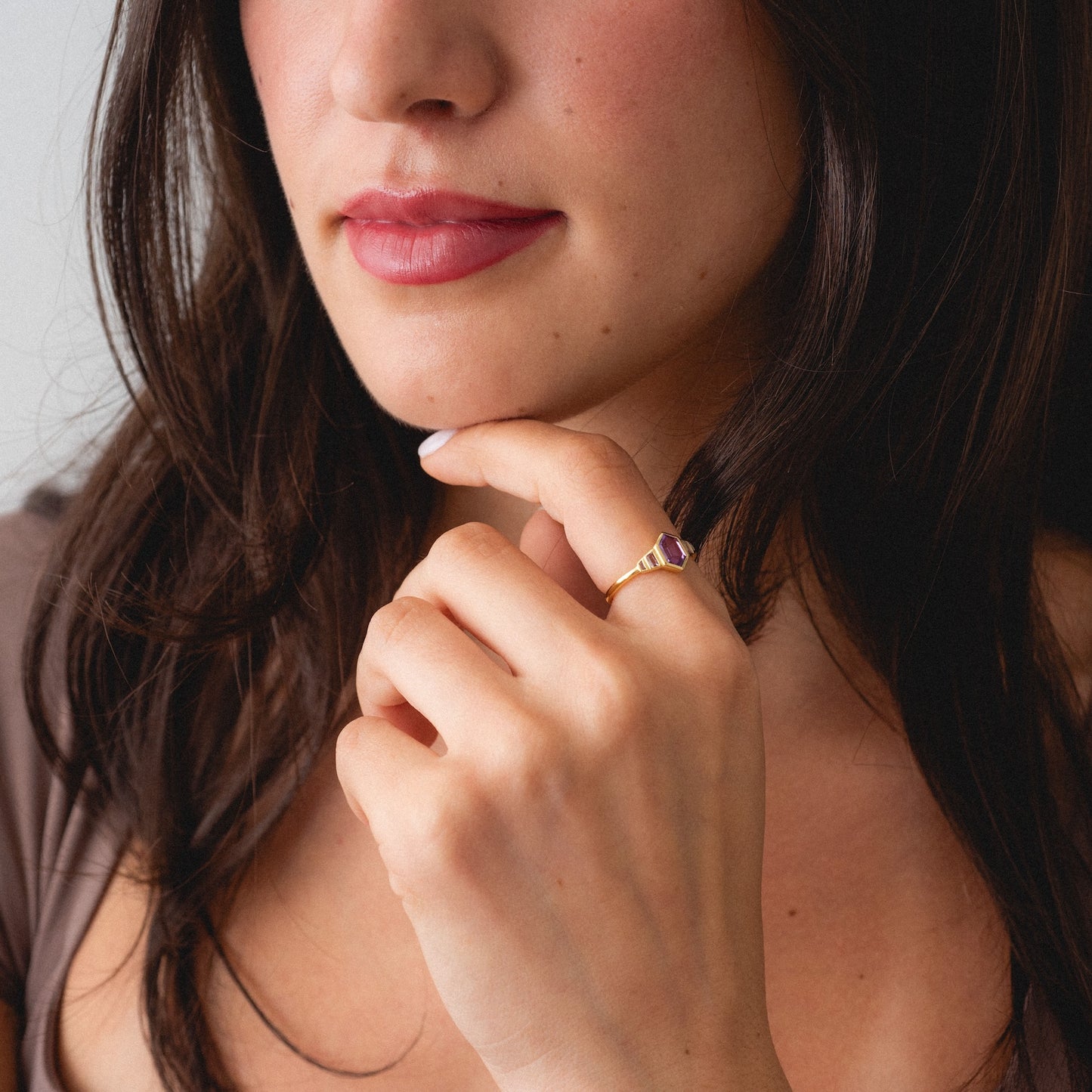 A woman with long dark hair touches her chin, showcasing the 2026 June Capsule Art Deco Alexandrite Ring on her finger. Only the lower half of her face and her hand are visible.