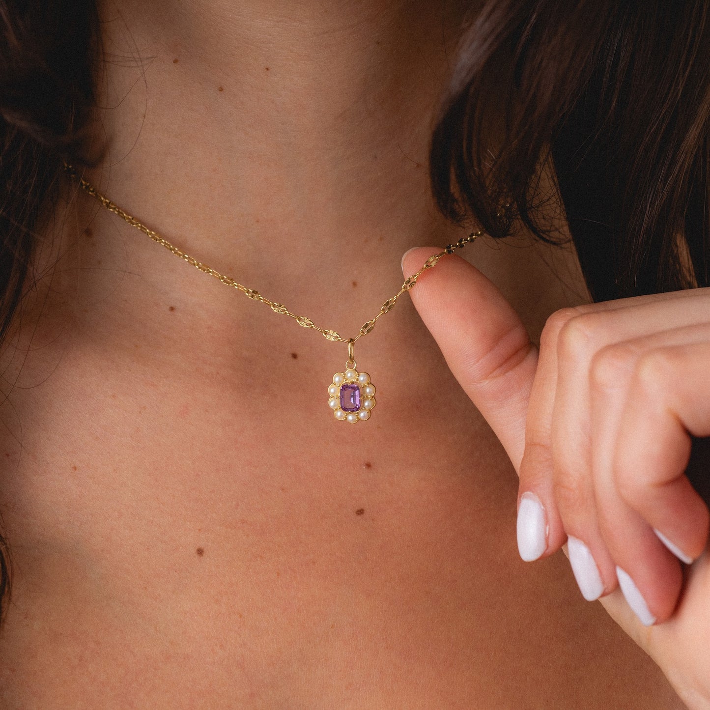 A person with fair skin and white nail polish touches the 2026 June Capsule Vintage Inspired Alexandrite and Pearl Charm, a gold necklace with a flower-shaped pendant featuring a pink stone center and small pearls.
