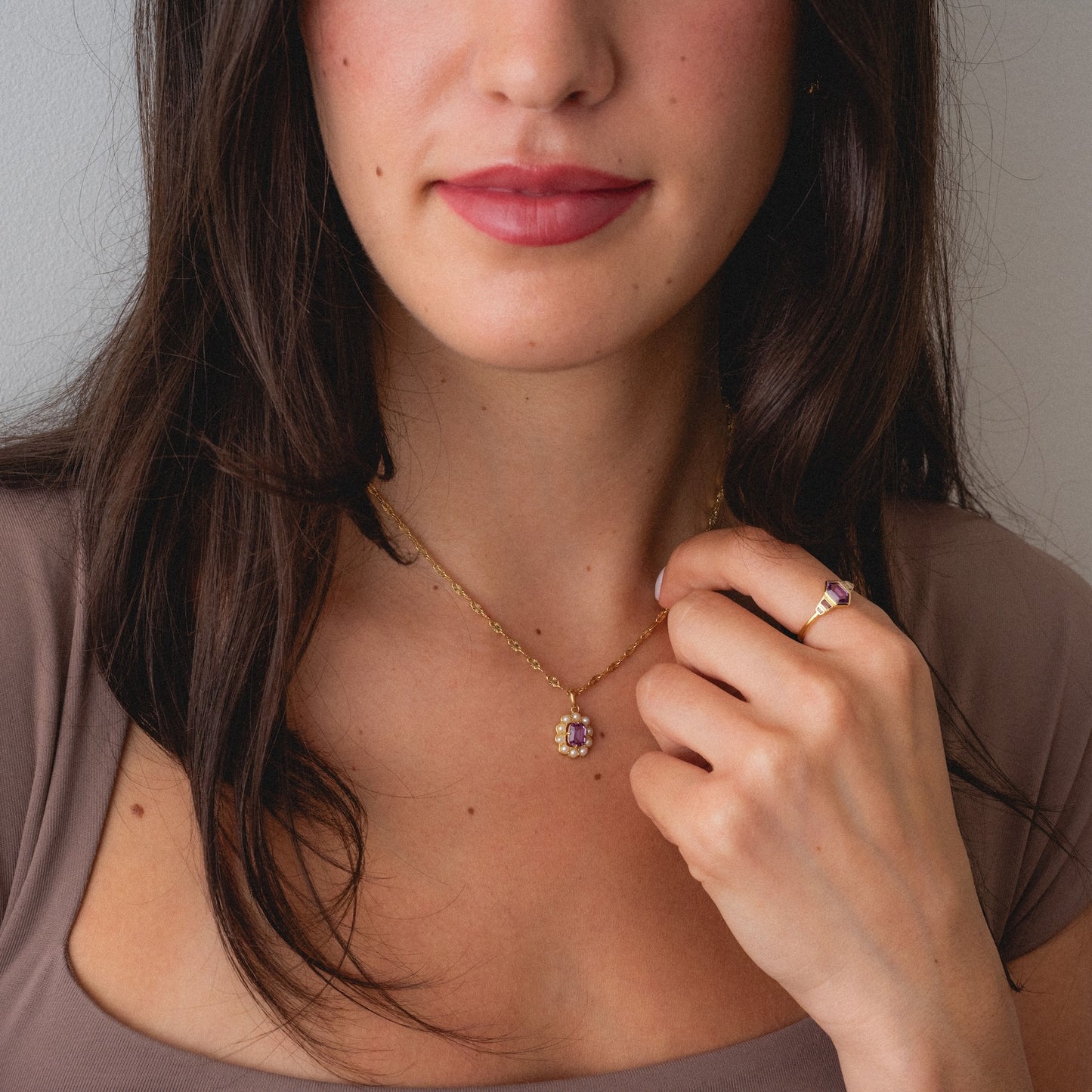 Woman with long dark hair wears the 2026 June Alexandrite and Pearl Capsule Set necklace and matching ring, featuring gold and purple stone details, holding the pendant charm near her collarbone. Set comes with a free gift.