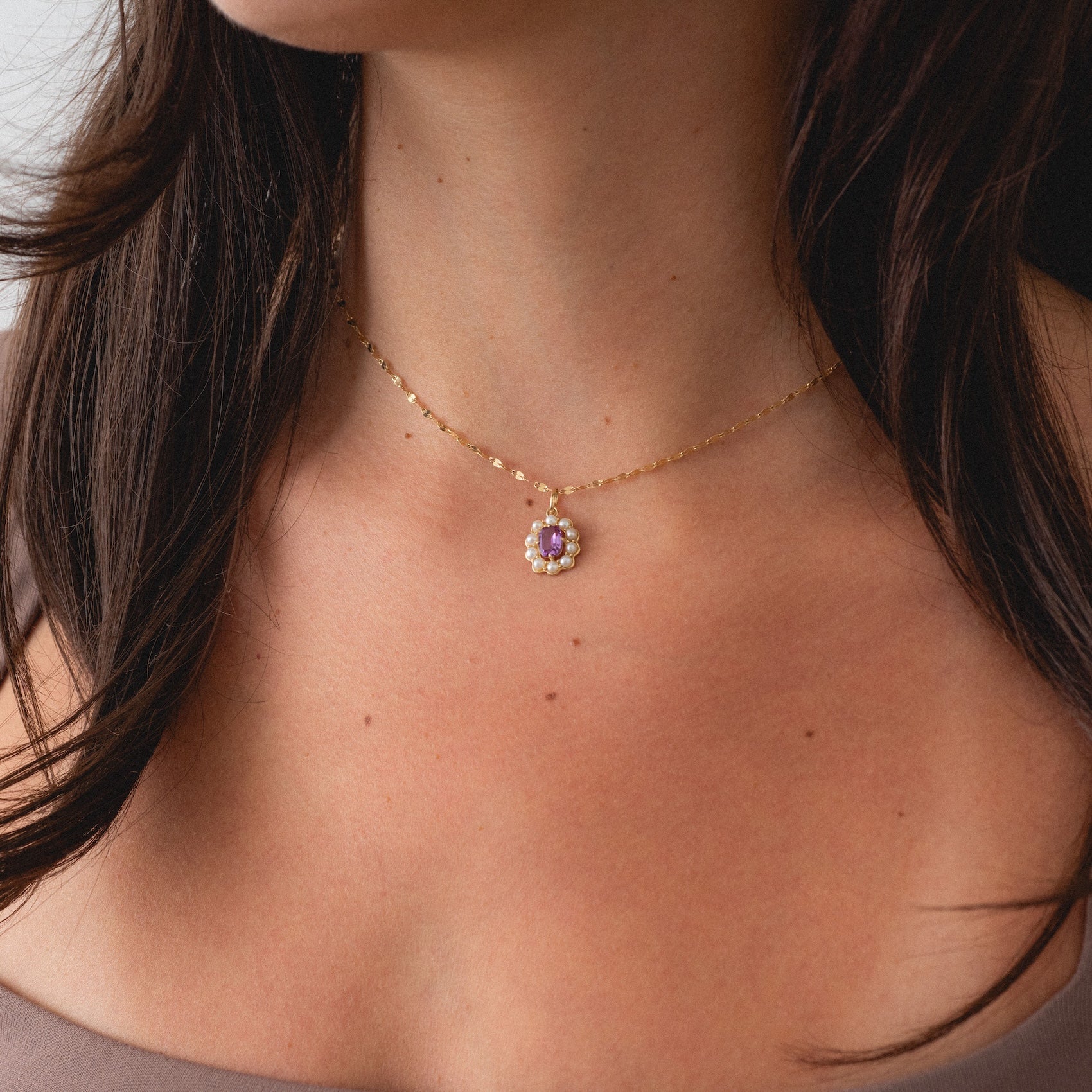 A woman wears the Solid Gold 2026 June Capsule Vintage Inspired Alexandrite and Pearl Charm necklace, featuring a delicate flower-shaped pendant. The focus is on her neck and collarbone, with her dark hair partially visible.