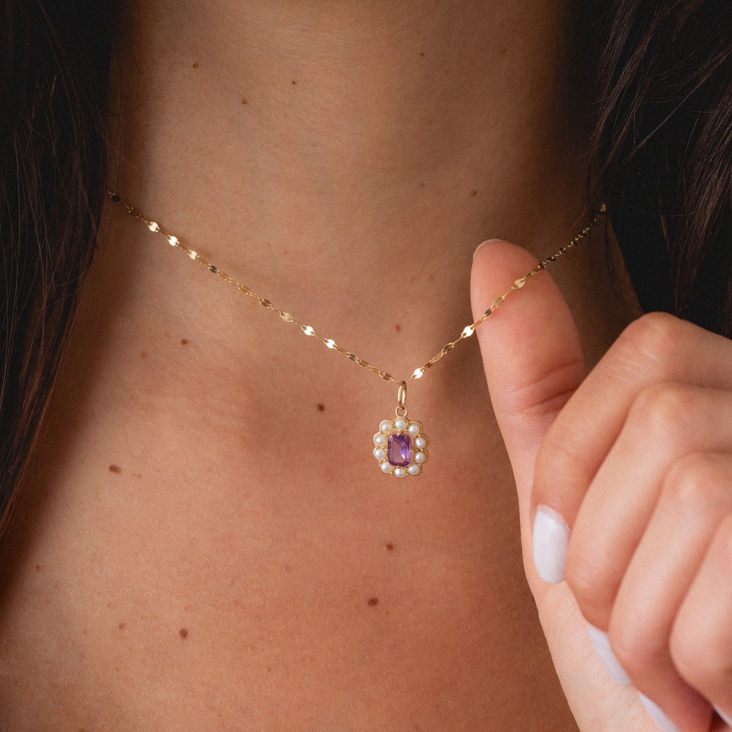 A close-up of a woman’s neck wearing the Solid Gold 2026 June Capsule Vintage Inspired Alexandrite and Pearl Charm as she gently holds its flower-shaped pendant, featuring a purple alexandrite center and pearl accents, between her fingers.