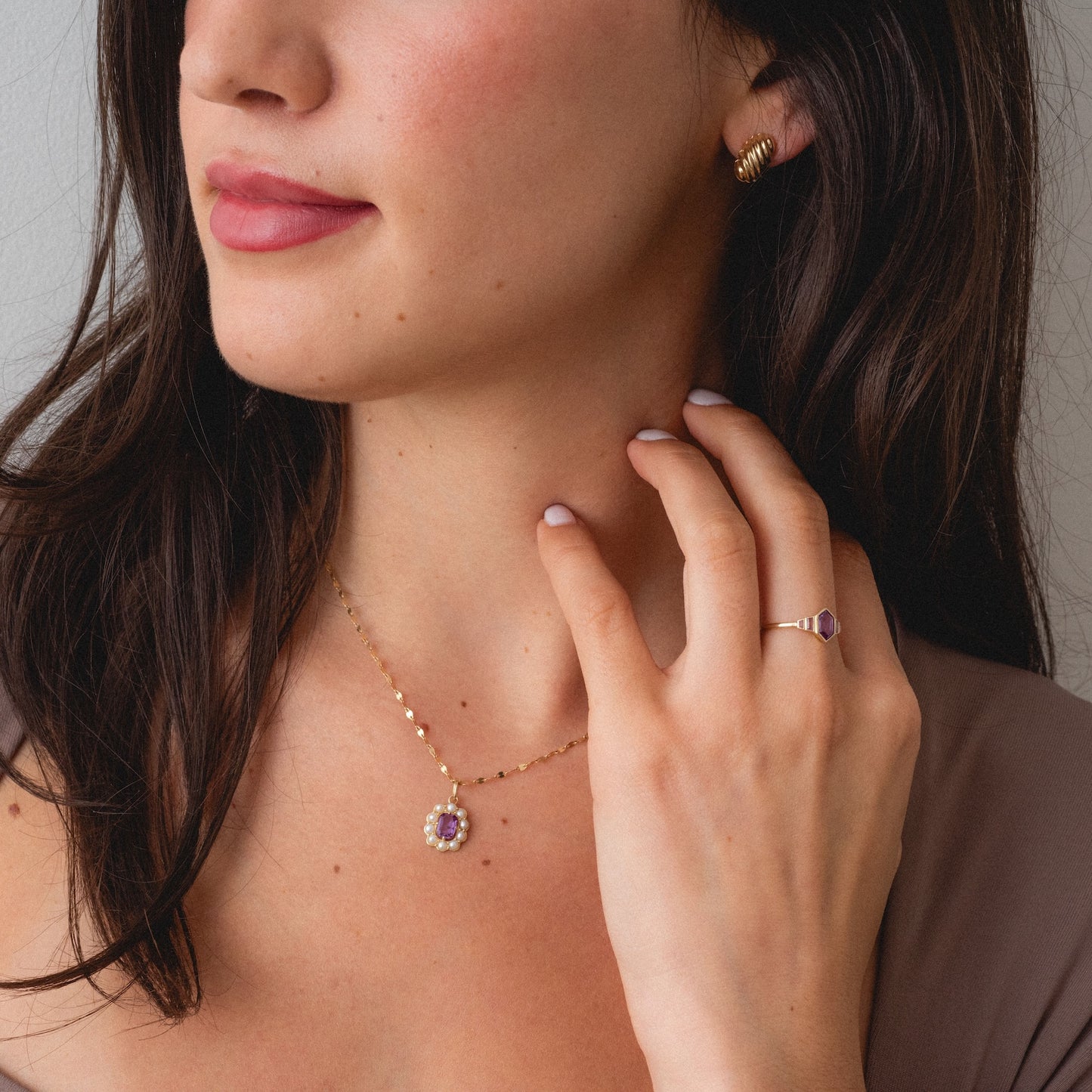 A woman with wavy dark hair wears the Solid Gold 2026 June Alexandrite and Pearl Capsule Set, featuring gold jewelry with purple Alexandrite stones. She touches her neck with manicured fingers and wears a taupe top. Free gift included.