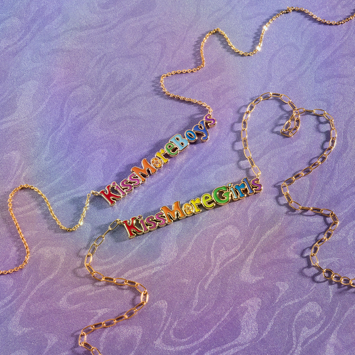 The Kiss More Girls Necklace features a gold chain and a playful, colorful "Kiss More Girls" pendant, shown lying on a purple textured surface.