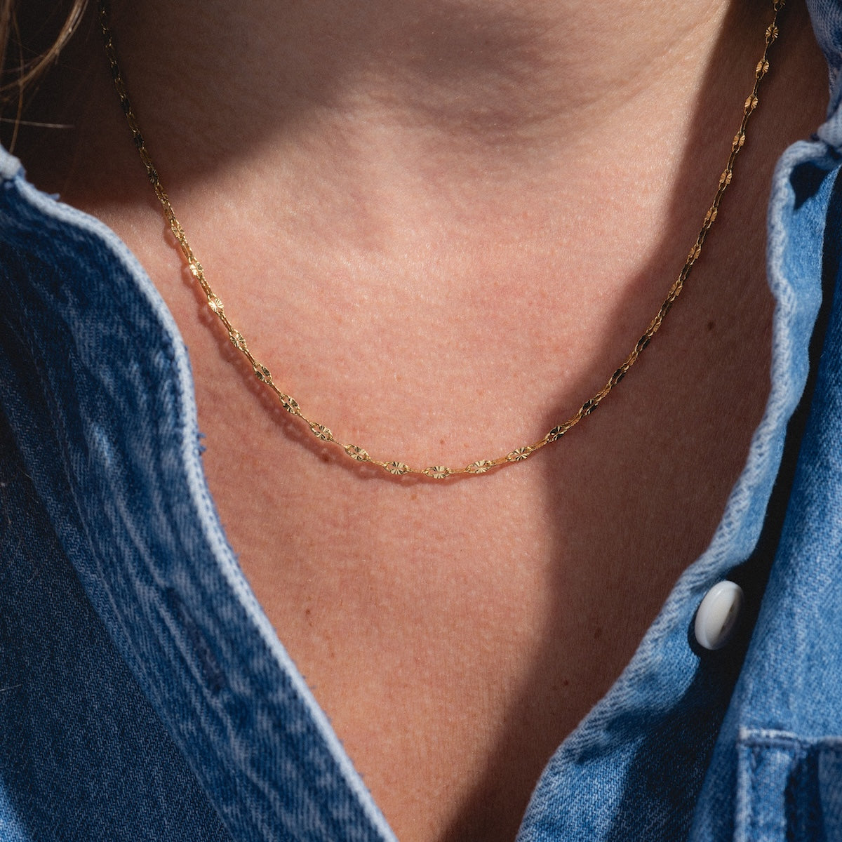 Close-up of a person wearing the Sparkle Link Chain, a delicate gold-plated necklace, paired with a blue denim shirt with white buttons and an open collar.