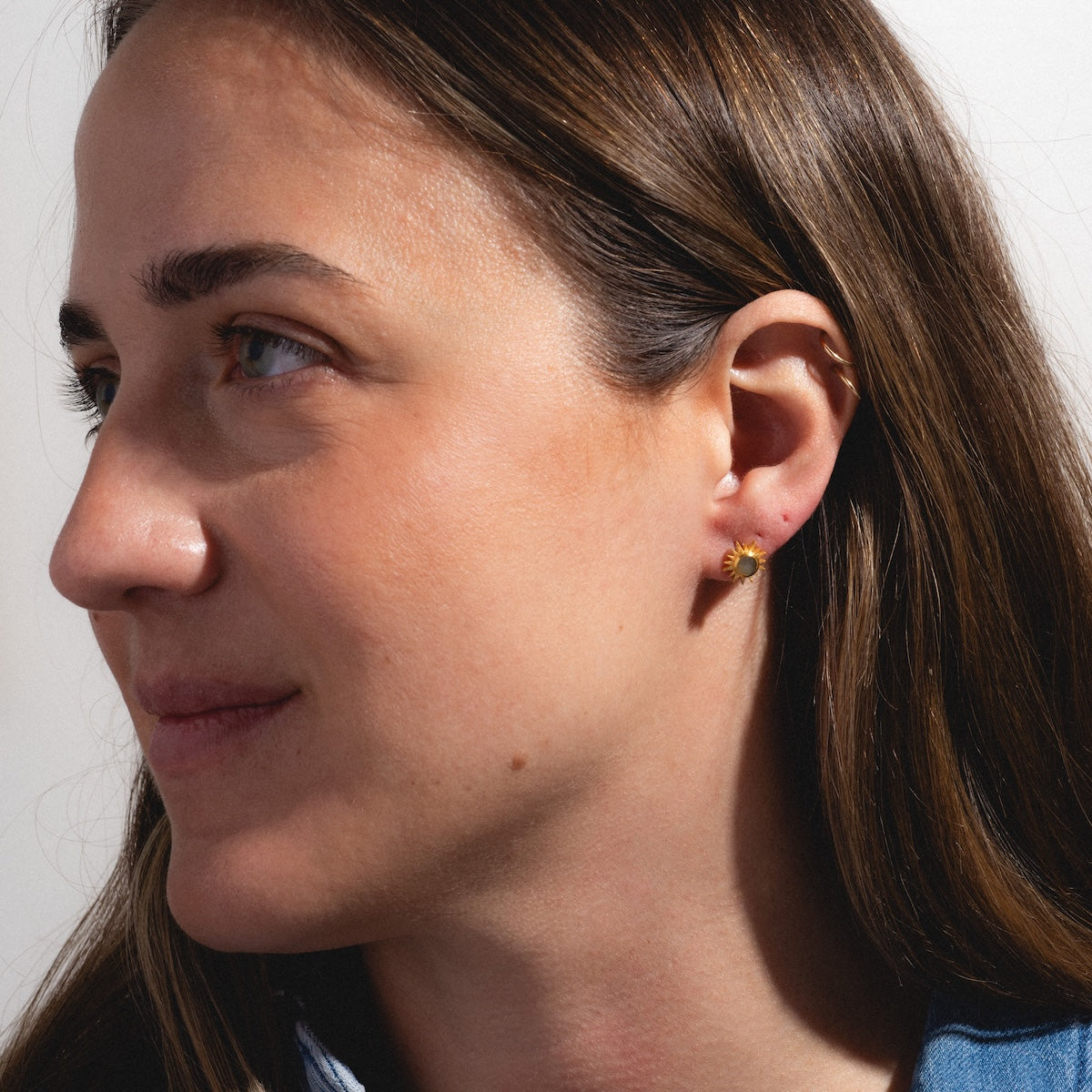 A woman with long brown hair looks to the left, wearing a Labradorite Sunburst Stud Earring by Satya Jewelry on her left ear. She has a calm expression in soft natural light against a plain background.