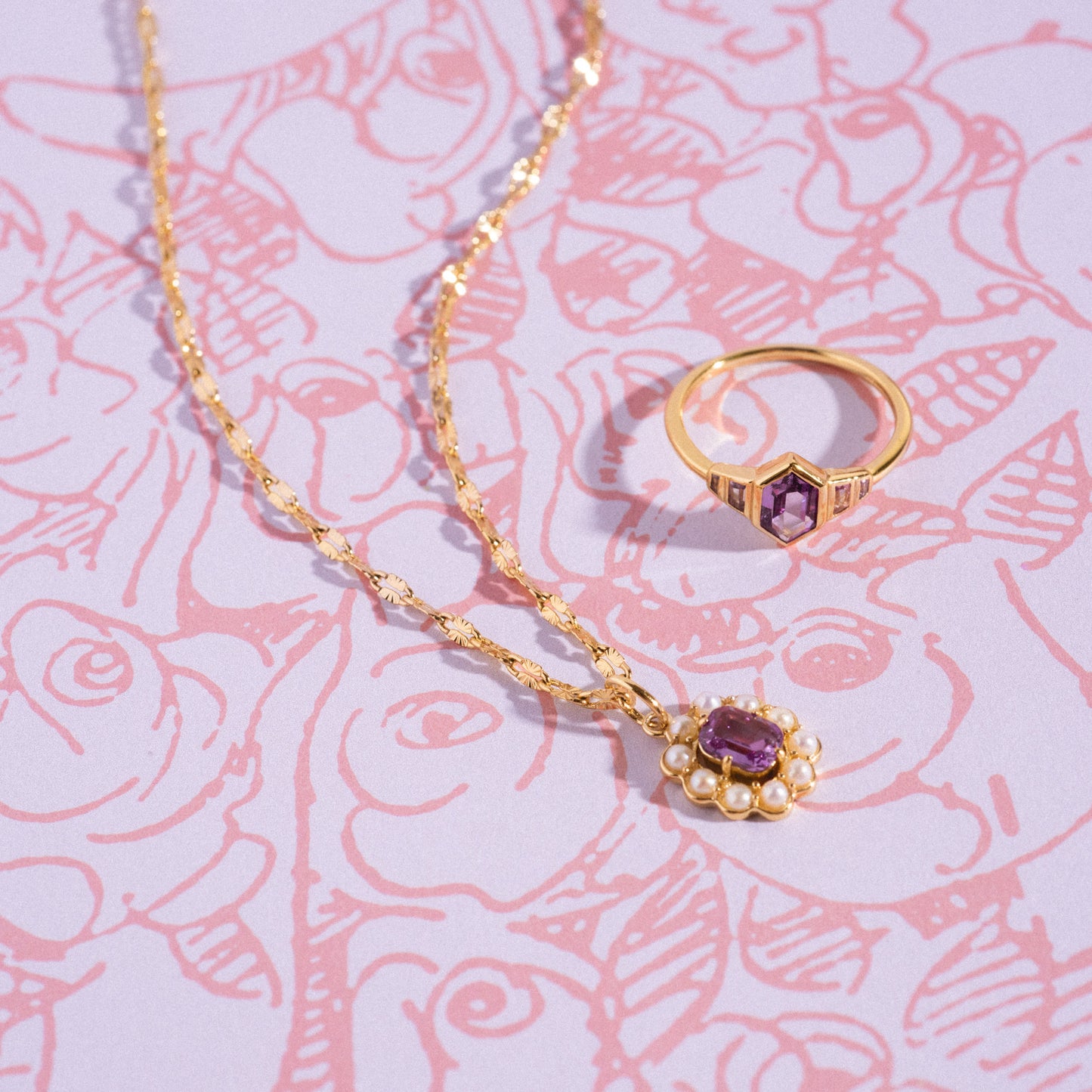 The 2026 June Capsule Art Deco Alexandrite Ring and a gold necklace with a purple gemstone pendant rest on a pink floral-patterned background.