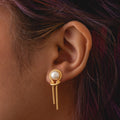 Close-up of a person's ear wearing Pearl Knot Earrings featuring a round white pearl and two delicate gold chain dangles. The person has brown hair with hints of purple.
