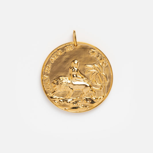 The Plated Zodiac Coin Charm Sample is a round yellow gold plated brass pendant, featuring a raised person riding a lion with palm trees. JULY 22 - AUG 23 and LEO are inscribed along the edges.
