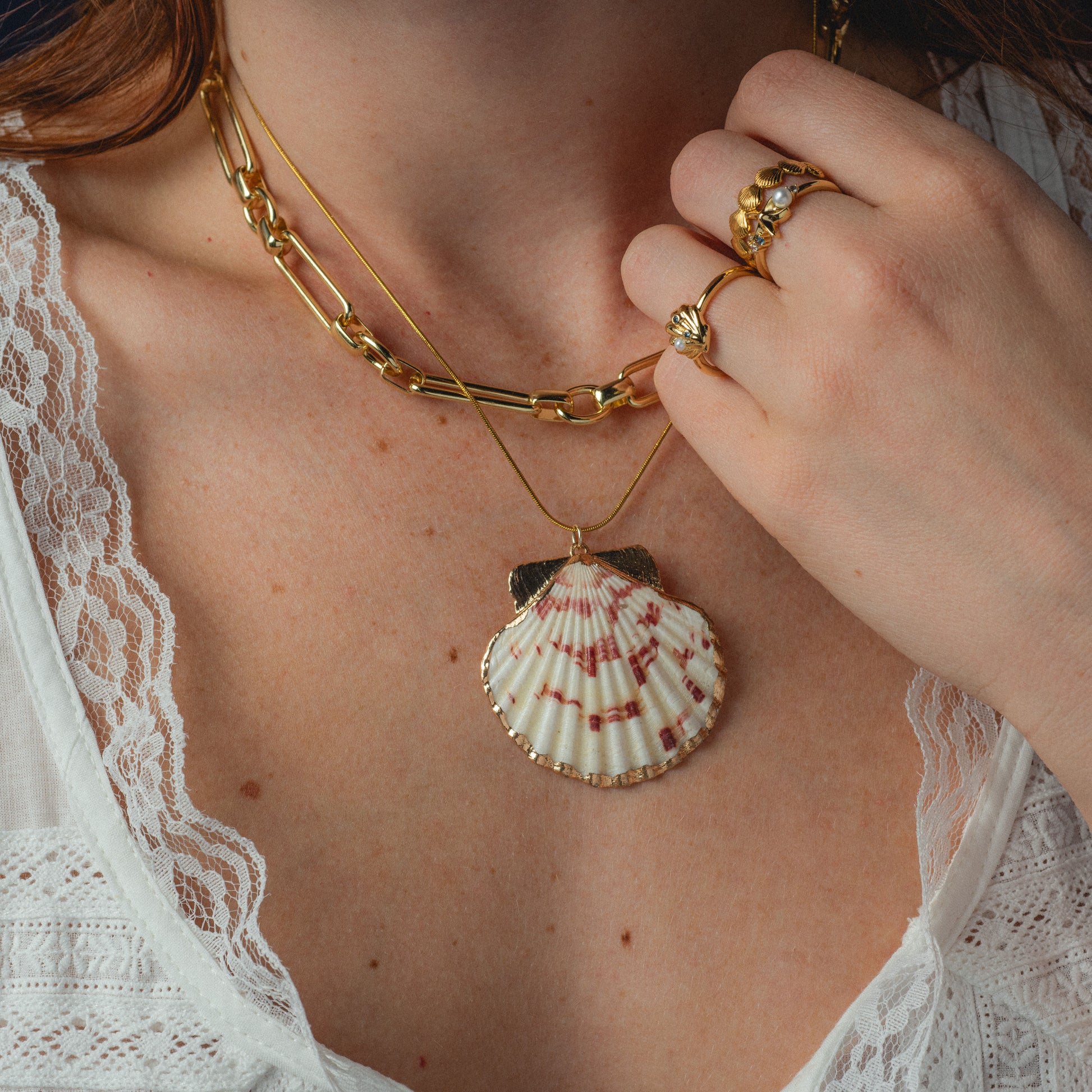 Person wearing a gold necklace with a shell pendant and a ring, against a neutral background.