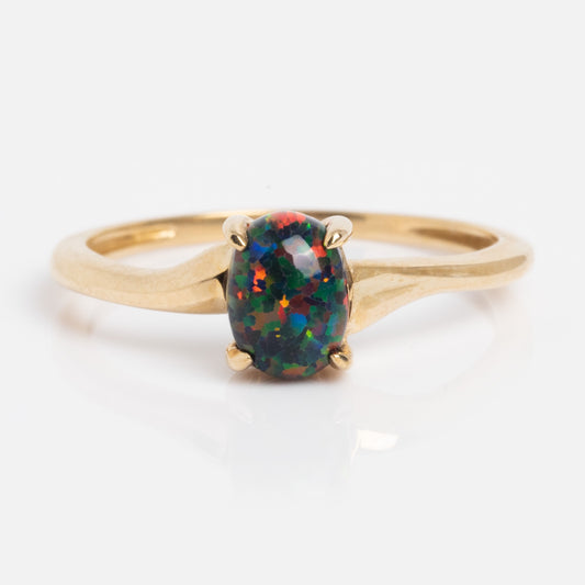 The Solid Gold Black Opal Oval Ring Size 7 Sample showcases a vibrant black opal with green, blue, and red specks in a four-prong setting on a smooth gold band, displayed against a white background.