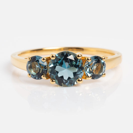 The London Blue Topaz Trilogy Ring Size 7 Sample is a gold ring with three round blue topaz stones—one large center stone and two smaller stones—displayed against a white background.