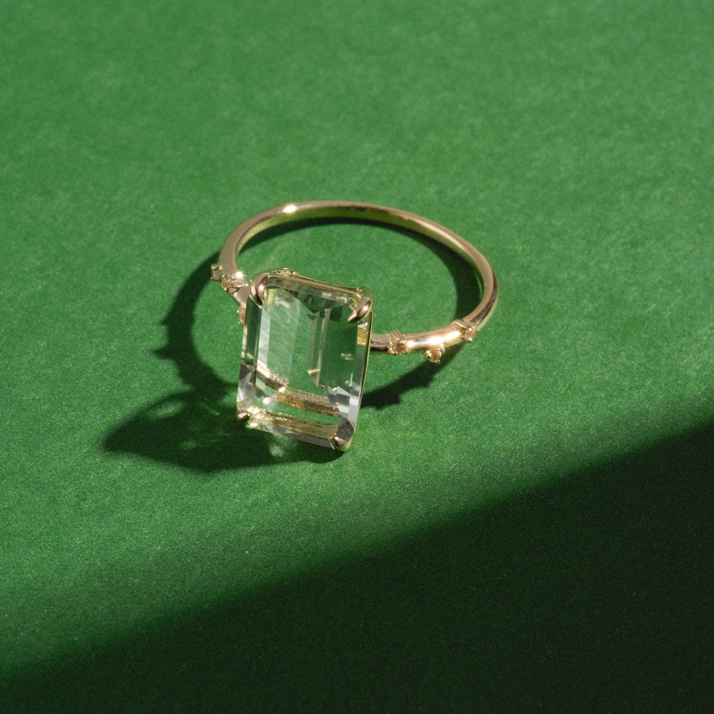 The Solid Gold Dewdrop Green Amethyst Statement Ring, with a striking rectangular green amethyst set in solid gold, is showcased on a textured green surface, its bold silhouette accented by a shadow over the bottom right corner.