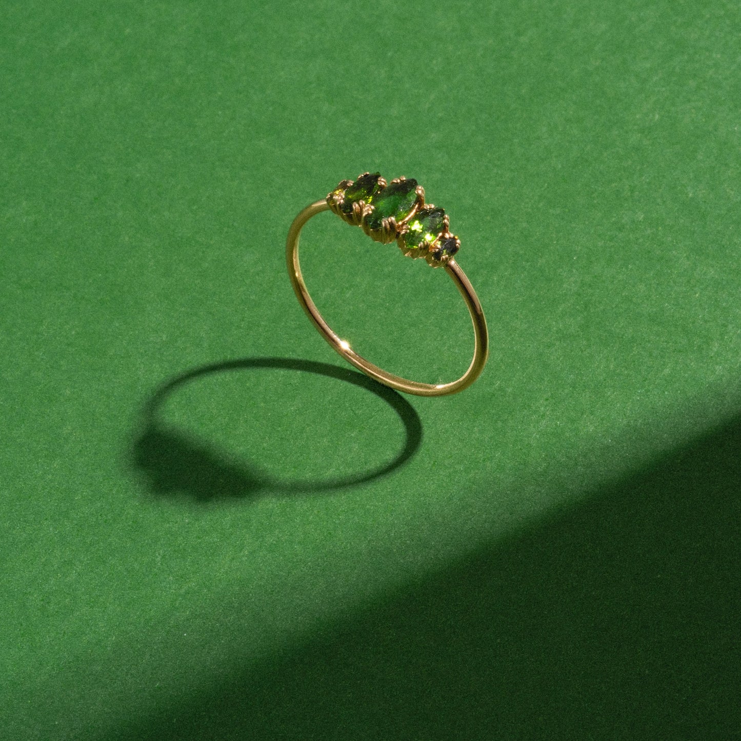 The Solid Gold Essential Marquise Cluster Ring, adorned with five small green gemstones, is displayed on a green surface and casts a distinct shadow to the left.