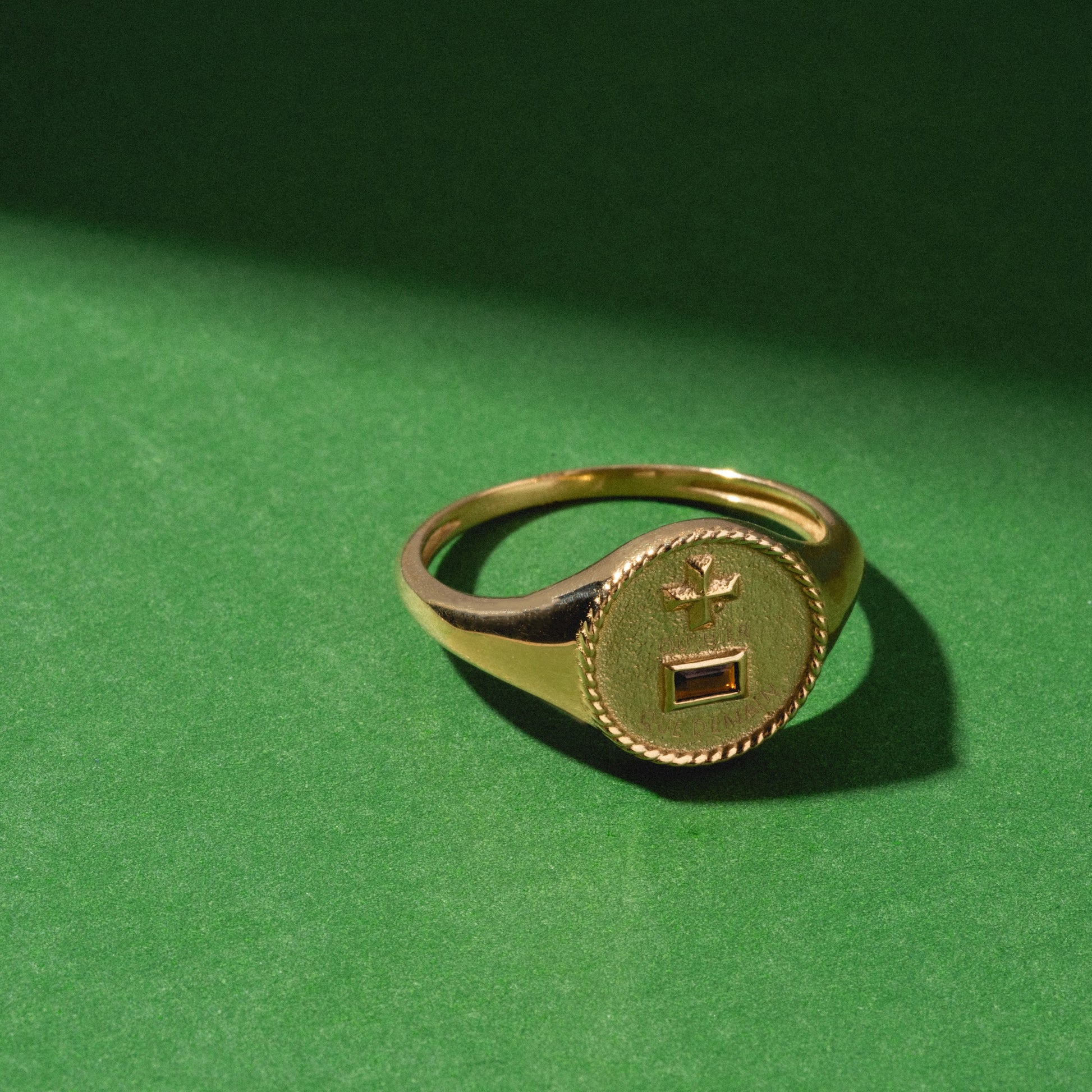 The Solid Gold Qu'hier Que Demain Signet Ring, with a round face and engraved design, rests on a green surface illuminated by angled sunlight.