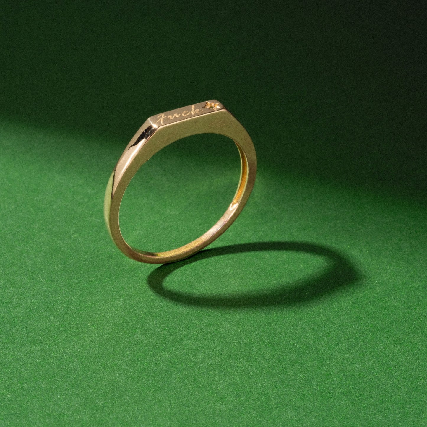 The Solid Gold Tiny Fuck Ring stands upright, engraved with “fuck” on top and casting a shadow on a green background—ideal for bold stacking rings with attitude.