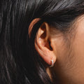 A close-up shows a person with long dark hair wearing the Dreaming About You Hoops—18k gold-plated hoop earrings featuring clear gemstones. The lighting highlights the sparkling jewelry and details of the ear.
