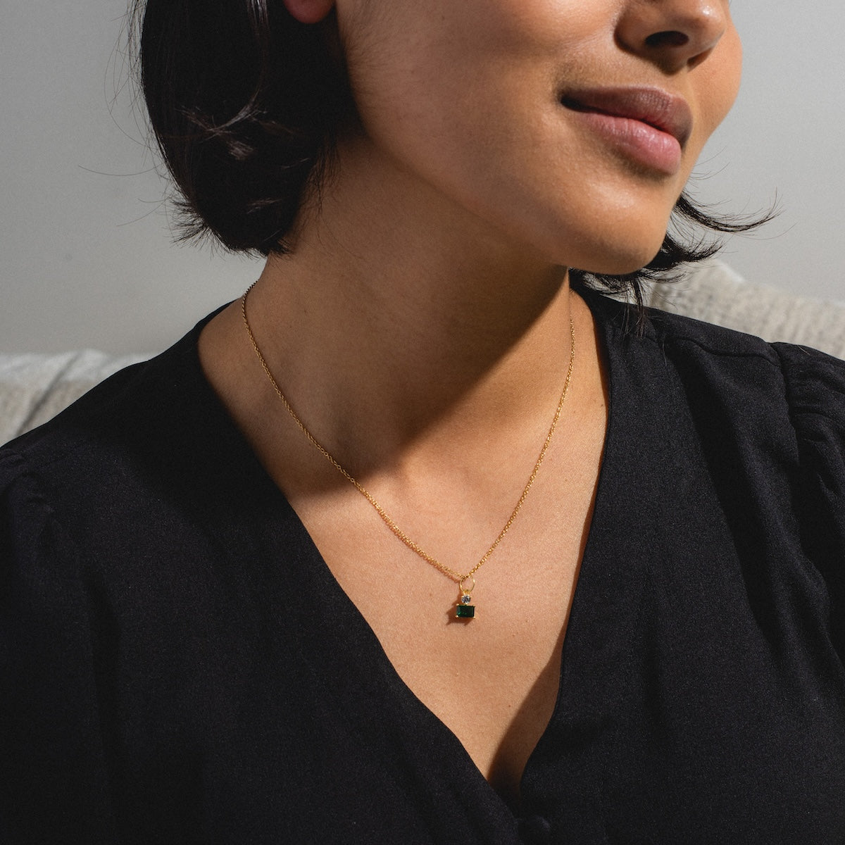 A woman in a black top wears the My Cherie Necklace—a gold plated chain with a delicate small emerald cubic zirconia pendant—while sitting in soft natural light.