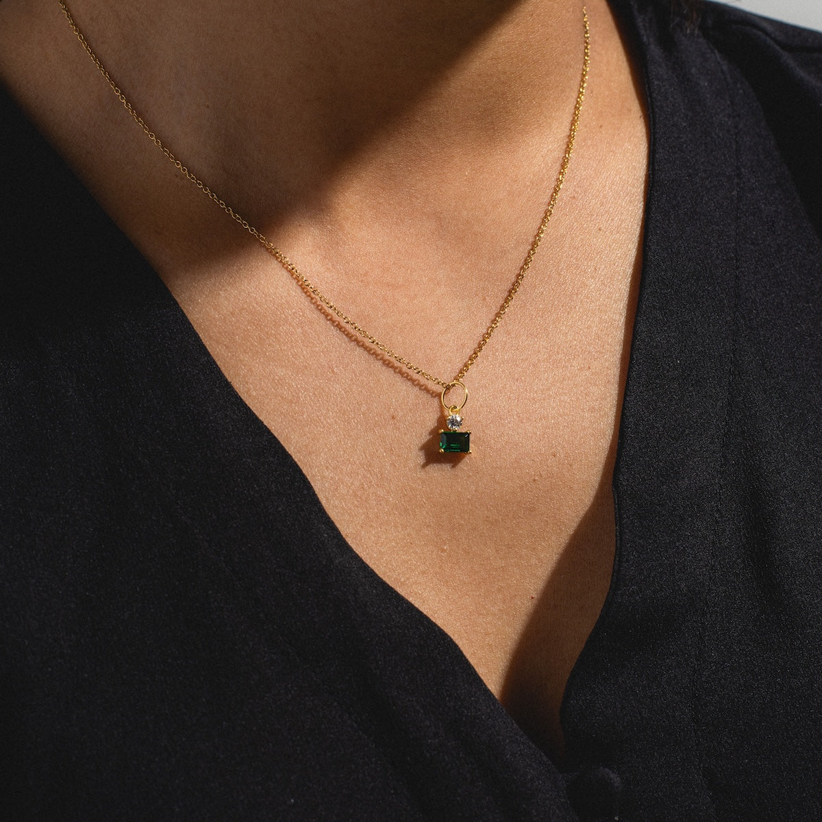 A person wearing the My Cherie Necklace—an emerald cubic zirconia with a rectangular green pendant on a delicate gold-plated chain—against their skin and a black V-neck top.