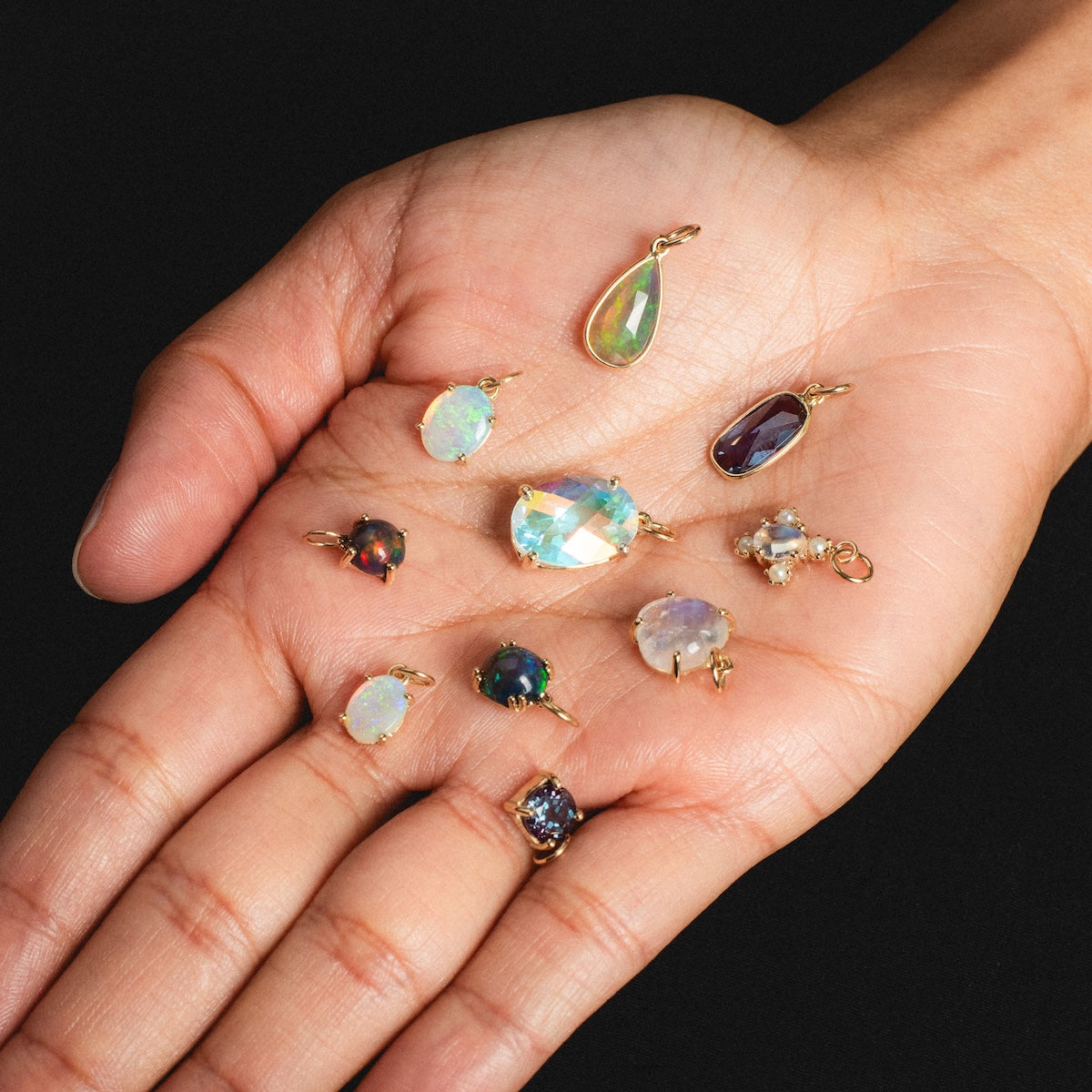 A hand shows nine unique, colorful gemstone pendants in 14k yellow gold against a black background. On the little finger is the One of a Kind 14k Elongated Pear Drop Black Welo Opal Charm worn as a ring.