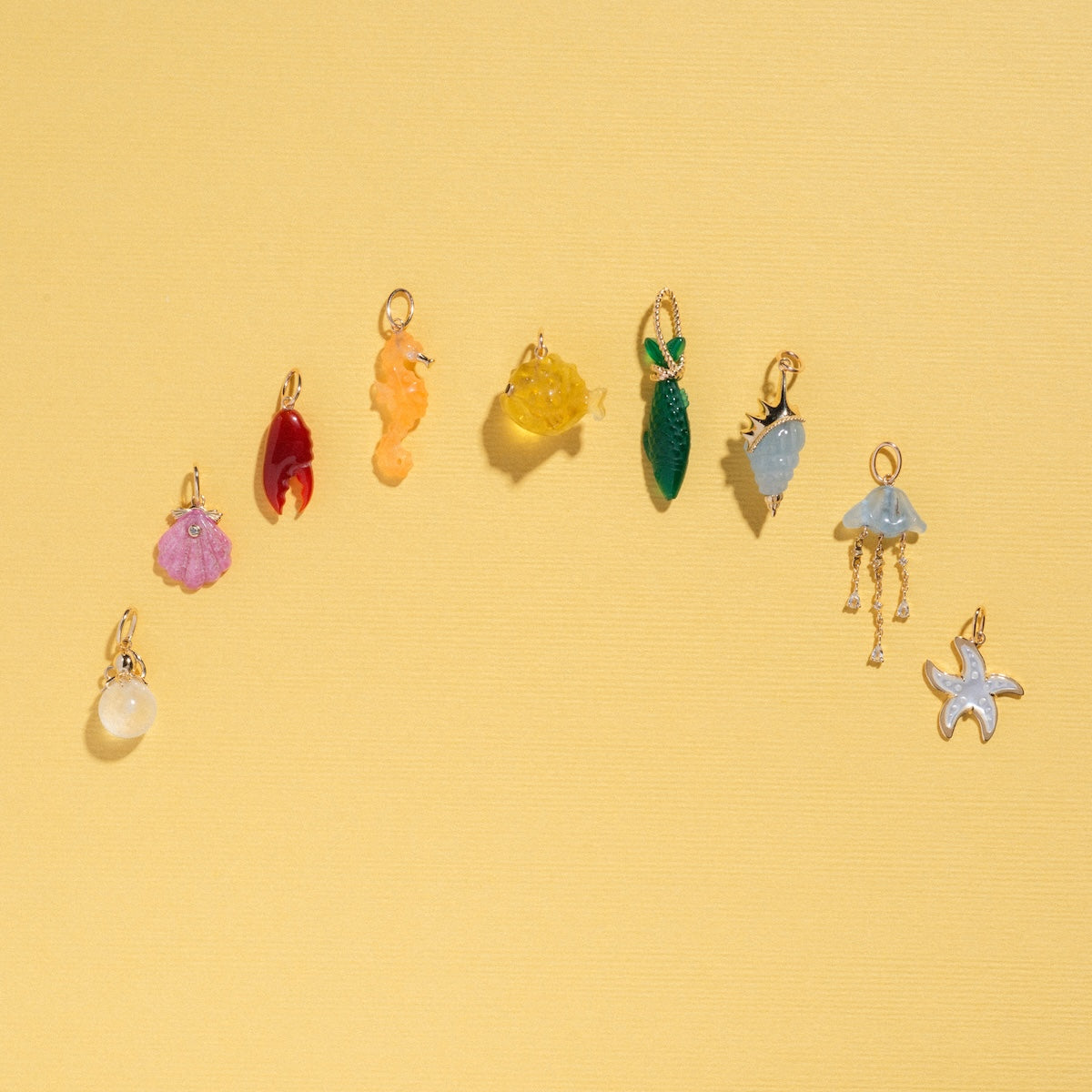 The Solid Gold Under The Sea Charm features nine vibrant sea-themed charms—including a shell, seahorse, spiral shell, and starfish—arranged in a curve on yellow, capturing the ocean’s enchanting beauty.