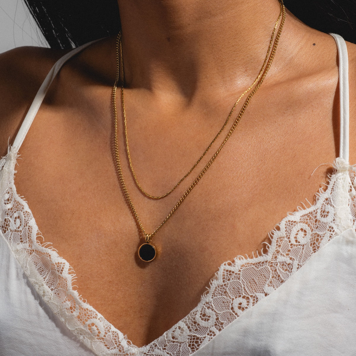 A person wearing a white lace-trimmed camisole and a layered necklace, including the Billie Necklace featuring a black coin pendant, stands against a neutral background.