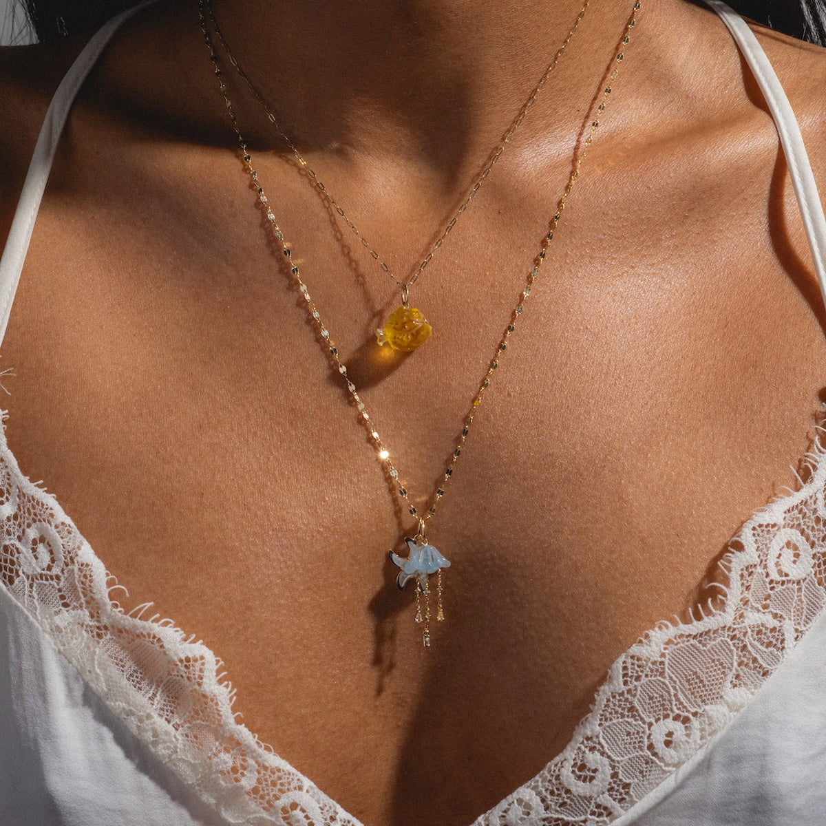 A close-up of a person wearing two delicate gold necklaces, including the Solid Gold Under The Sea Charm with a blue pendant, layered over a white lace-trimmed top, creating an effortless, radiant look.