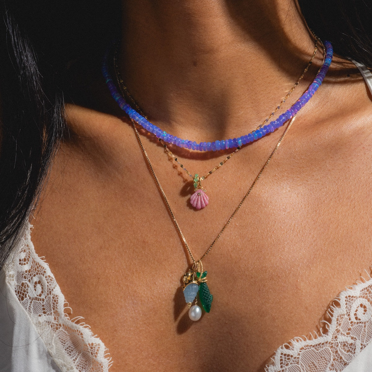 A close-up of layered necklaces: a blue beaded choker, gold chain with the Solid Gold Under The Sea Charm—featuring pink seashell, leaf, pearl, and green details—adds siren glow to their beauty over a white lace-trimmed top.