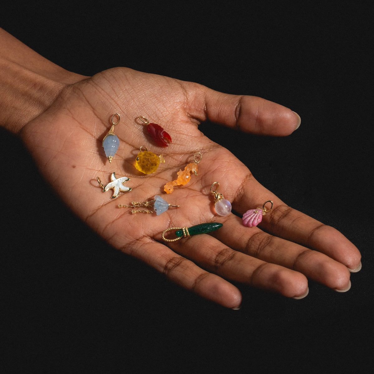 A hand against a black background displays the Solid Gold Under The Sea Charm, glowing with vibrant stones, shells, a bird, and colorful beads in blue, yellow, pink, green, and red.