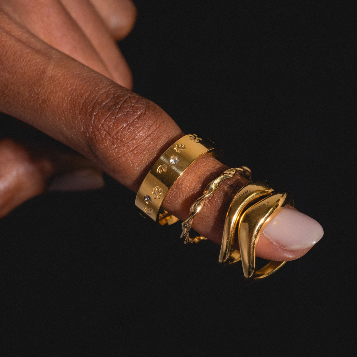 A hand with medium brown skin displays a stack of four Souvenirs Rings in yellow gold, including a twisted band, each with unique designs. Set against a black background, the natural and glossy thumb nail complements the look.