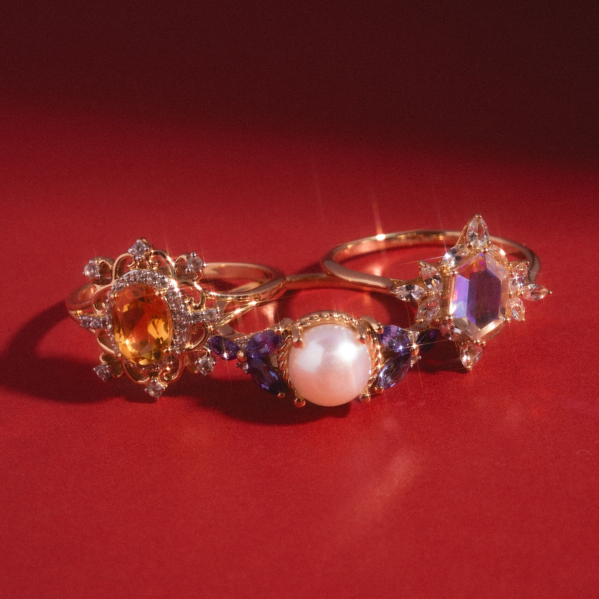 The 14k Angel Aura Quartz Splendor Statement Ring shines alongside two ornate solid gold rings: one with a yellow gemstone and another with a white sapphire cluster and pearl, all sparkling on a red surface.