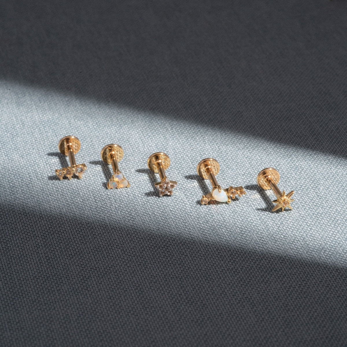 Five Solid Gold Starburst Flat Back Studs with different shapes—flower, heart, star, another heart, and starburst—are arranged in a row on a gray textured surface with a diagonal beam of light across them.