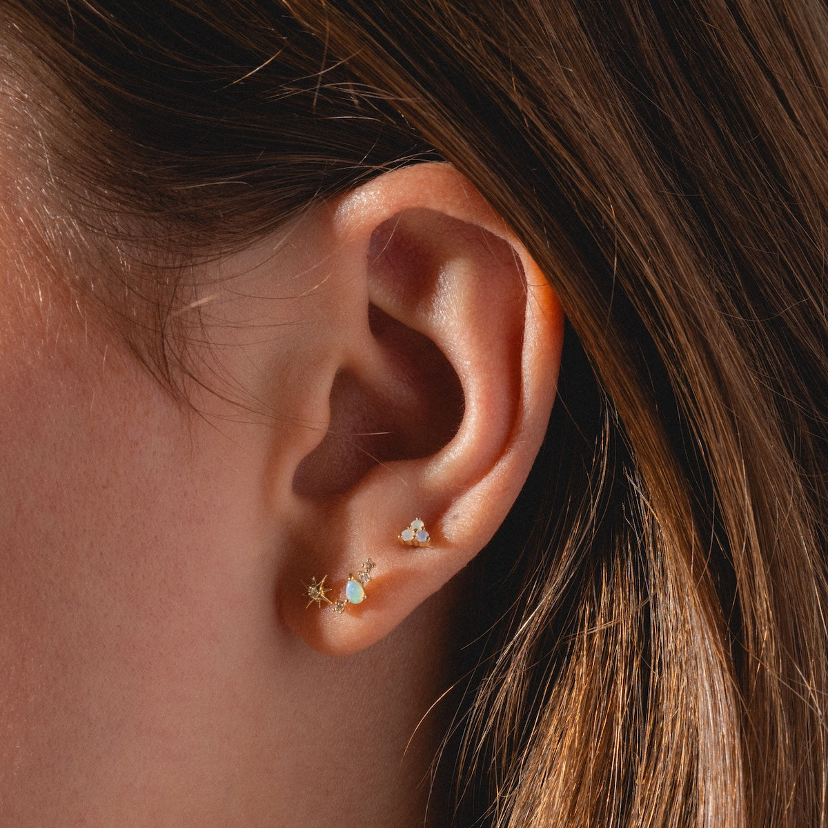 Close-up of an ear adorned with Solid Gold Starburst Flat Back Studs, including a small star-shaped opal stud, a tiny round stud, and a delicate bee-shaped stud. Brown hair frames the ear.