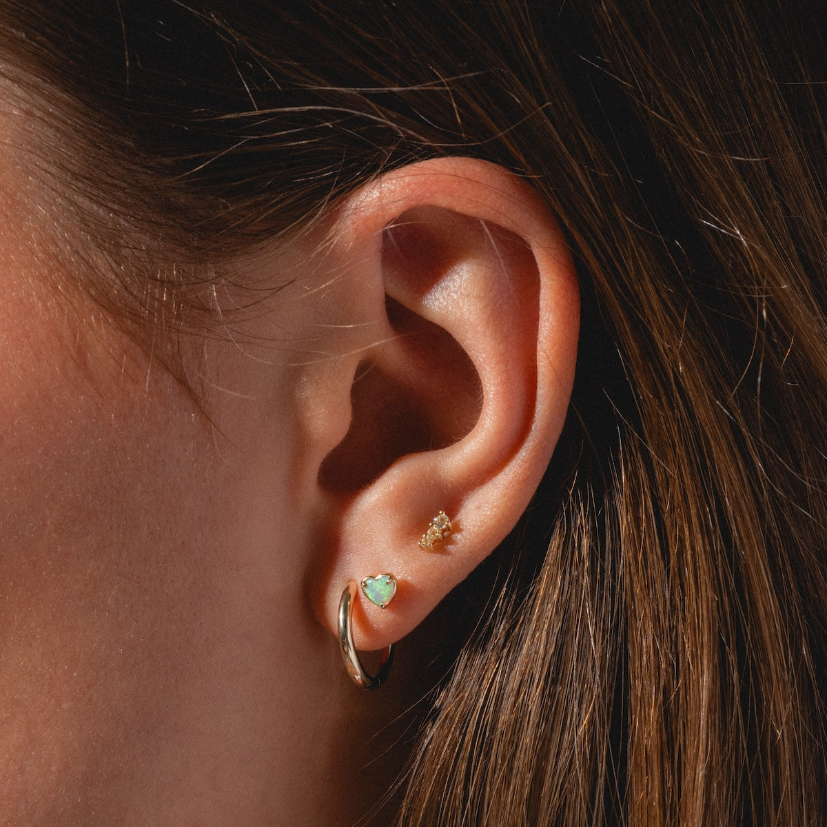 Close-up of a person's ear with three piercings: a gold hoop in the lobe, a turquoise heart stud above, and the Solid Gold Tapered Shimmer Flat Back Stud higher up. The person has straight brown hair.