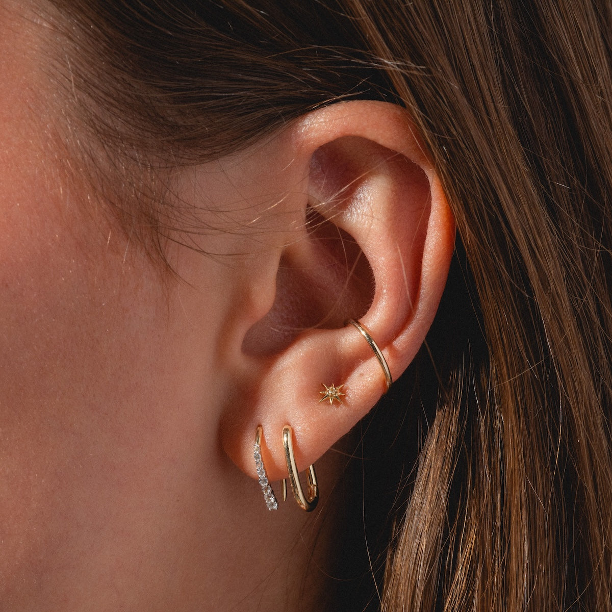 A close-up of an ear with multiple gold earrings, including small hoops and a Solid Gold Starburst Flat Back Stud, set against brown hair.