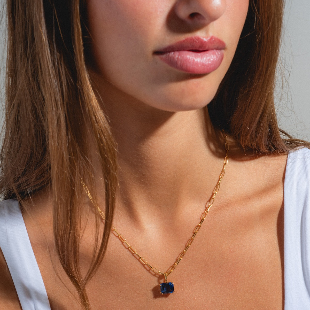 A woman with long brown hair wears a white top and the Born to Shine Birthstone Necklace, featuring a blue gemstone pendant on a gold chain. Soft natural lighting highlights her neck, lips, and the personalized necklace.