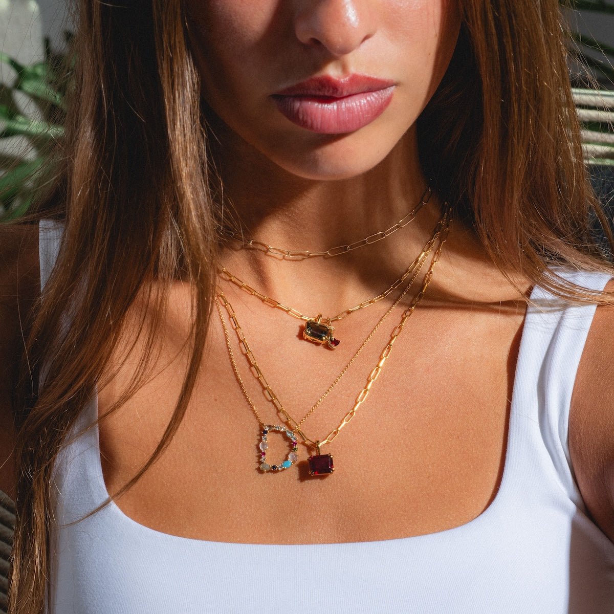 A woman in a white top wears three gold chains, including a paperclip chain and the Born to Shine Birthstone Necklace, layered elegantly. Sunlight highlights her skin and jewelry against a softly blurred background of green plants.