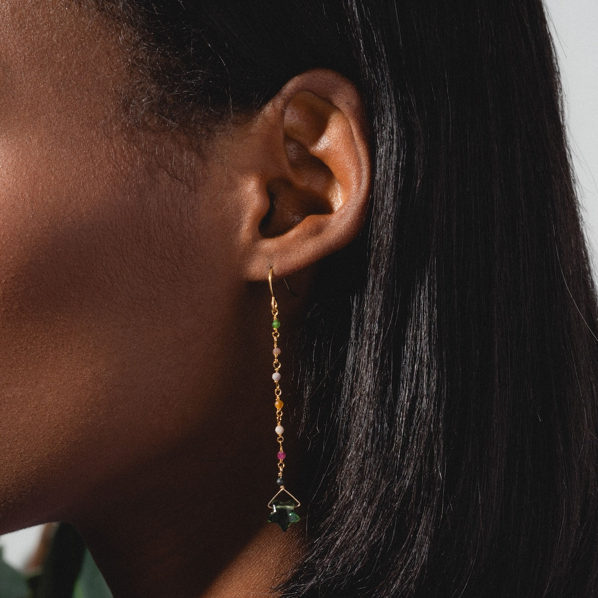 Close-up of a person wearing Bangalore Earrings featuring colorful beads, a green star-shaped charm, and yellow gold plating. Their straight, dark hair contrasts with the neutral background.
