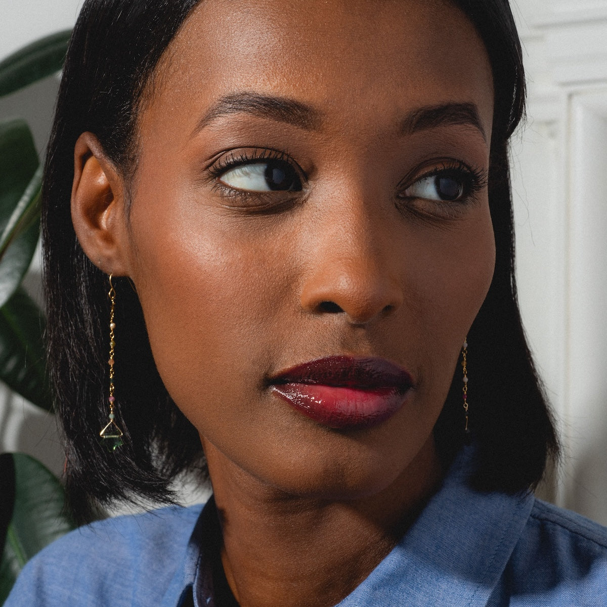 A woman with smooth, dark skin and straight black hair looks to the side, wearing bold dark red lipstick and yellow gold plated Bangalore Earrings. She pairs them with a blue collared shirt; a leafy plant appears in the background.