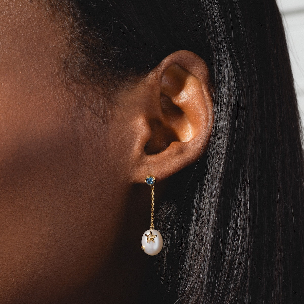 Close-up of a womans ear with straight black hair, wearing Gem Accent Pearl Drop Earrings—14k gold plated with a blue gem stud, delicate chain, and a white pearl drop pendant adorned with a small gold star.