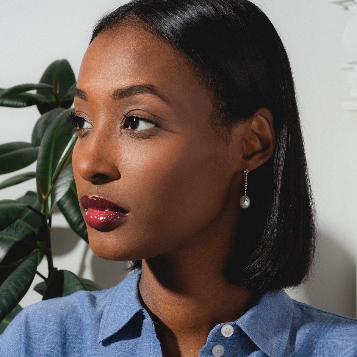 A woman with straight, shoulder-length dark hair wears a blue collared shirt and a single Gem Accent Pearl Drop Earring, looking to the left. A green plant is in the background, with soft light casting shadows on the wall.