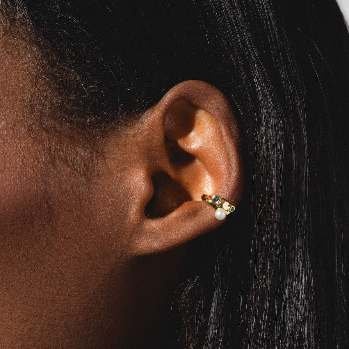 A close-up highlights a gold hoop earring with pearl and gemstone, paired with the delicate Gem Droplet Ear Cuff on smooth, dark skin and straight, dark hair.