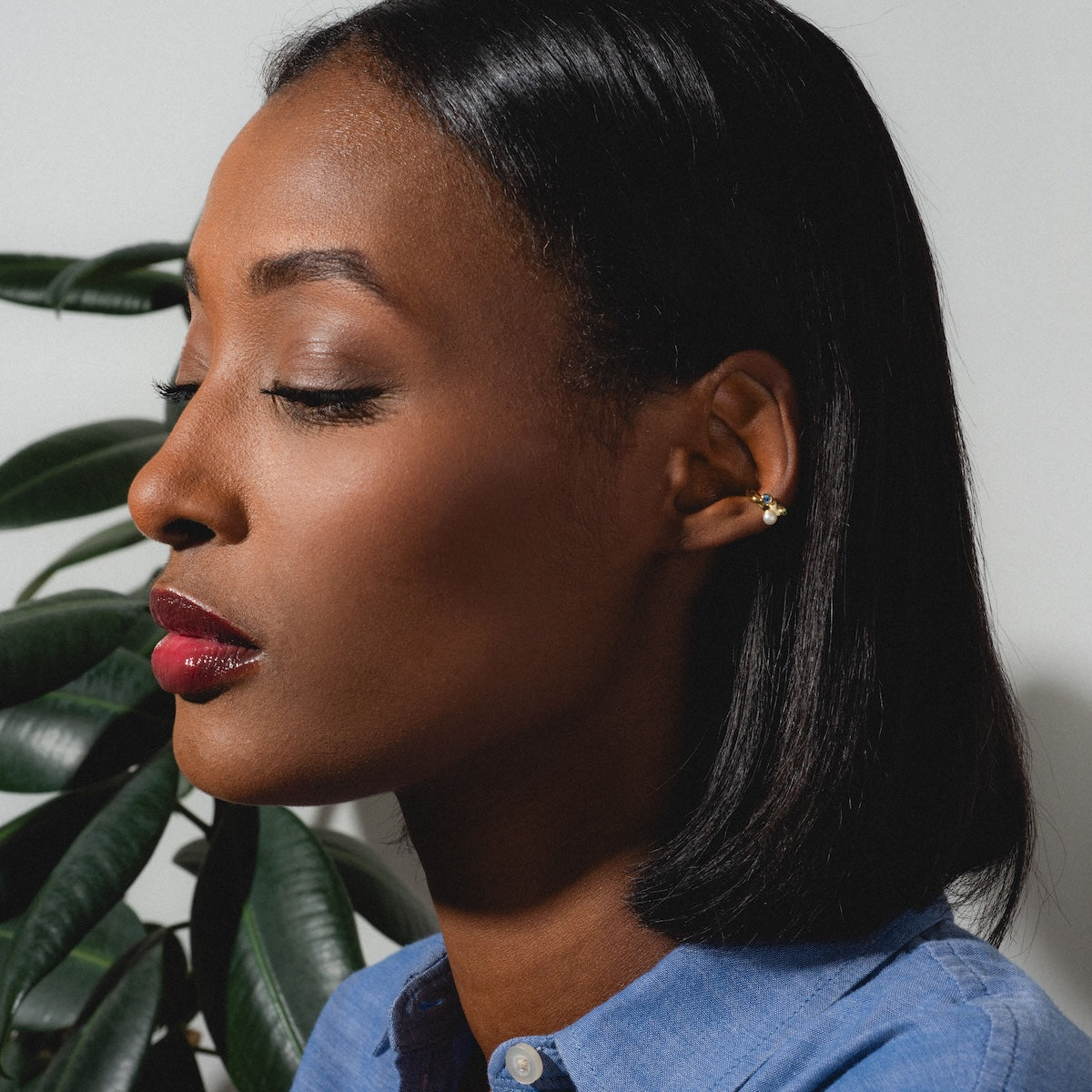 A woman with closed eyes and short black hair, wearing a blue shirt and the Gem Droplet Ear Cuff, poses in profile against a white background with green plant leaves behind her.