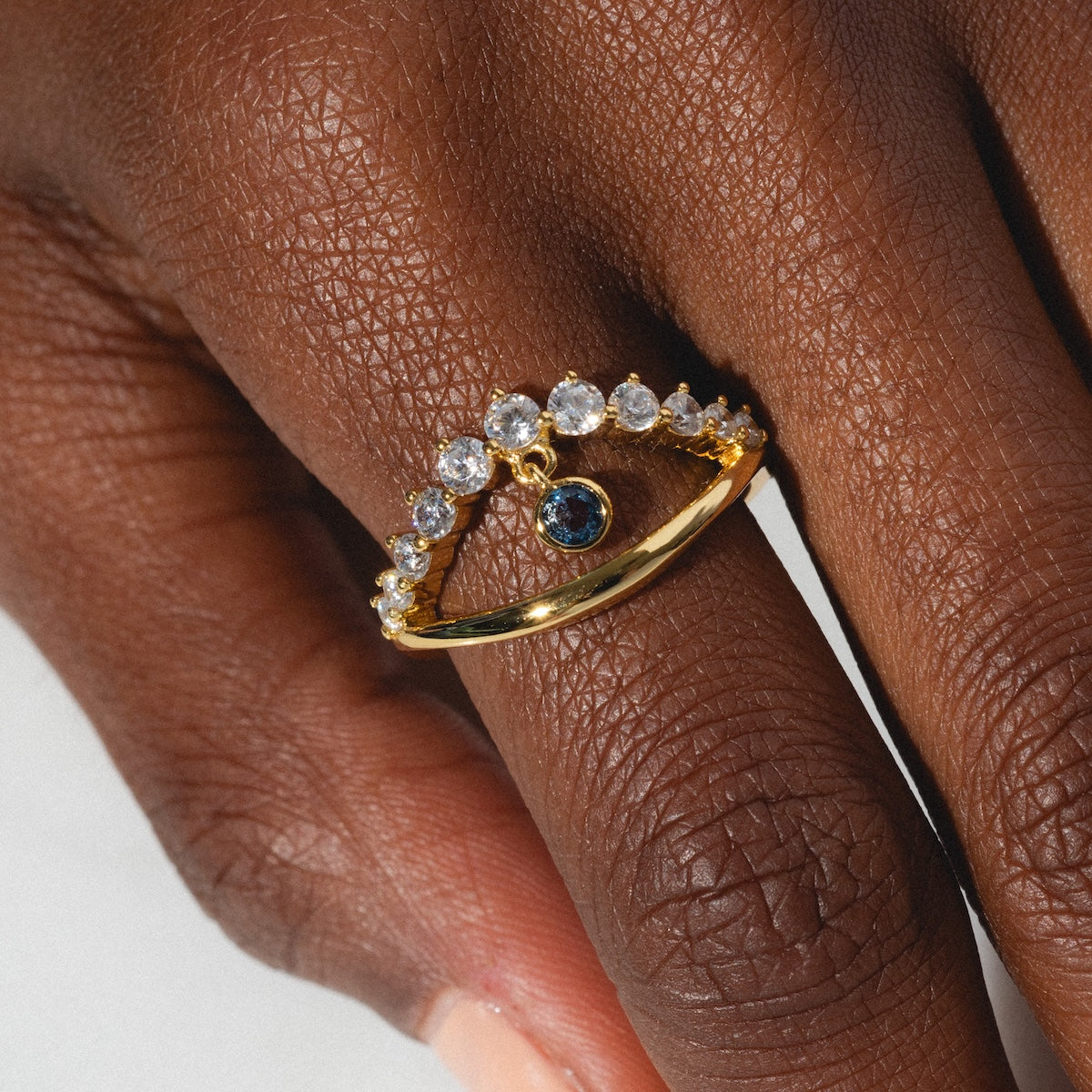 A close-up of a hand wearing the Mystique Evil Eye Charm Ring, a 14k yellow gold plated piece featuring sparkling gems and a central blue stone in an elegant curved design.