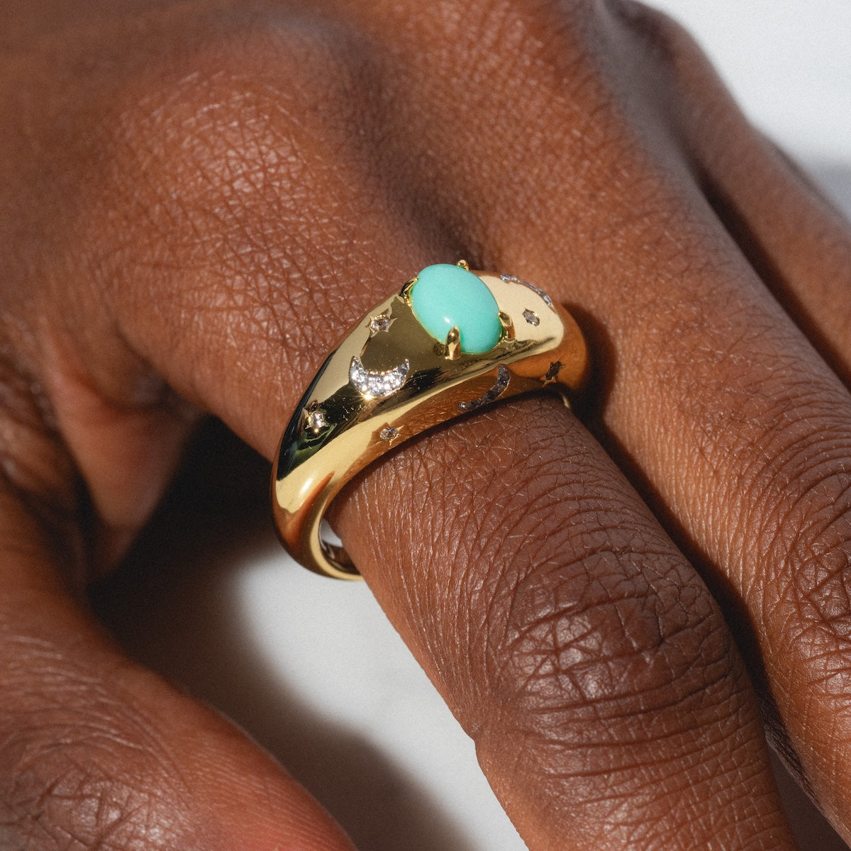 A hand displays the Turquoise Sky Dome Ring—gold-plated, featuring an oval green gemstone and small clear stones—set against a neutral background.