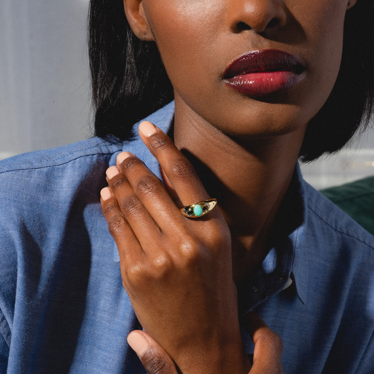 A woman in a blue shirt rests her hand on her chest, displaying the Turquoise Sky Dome Ring with a large green stone. Her glossy dark red lips and the ring are the focus of the cropped photo showing her lower face and hand.