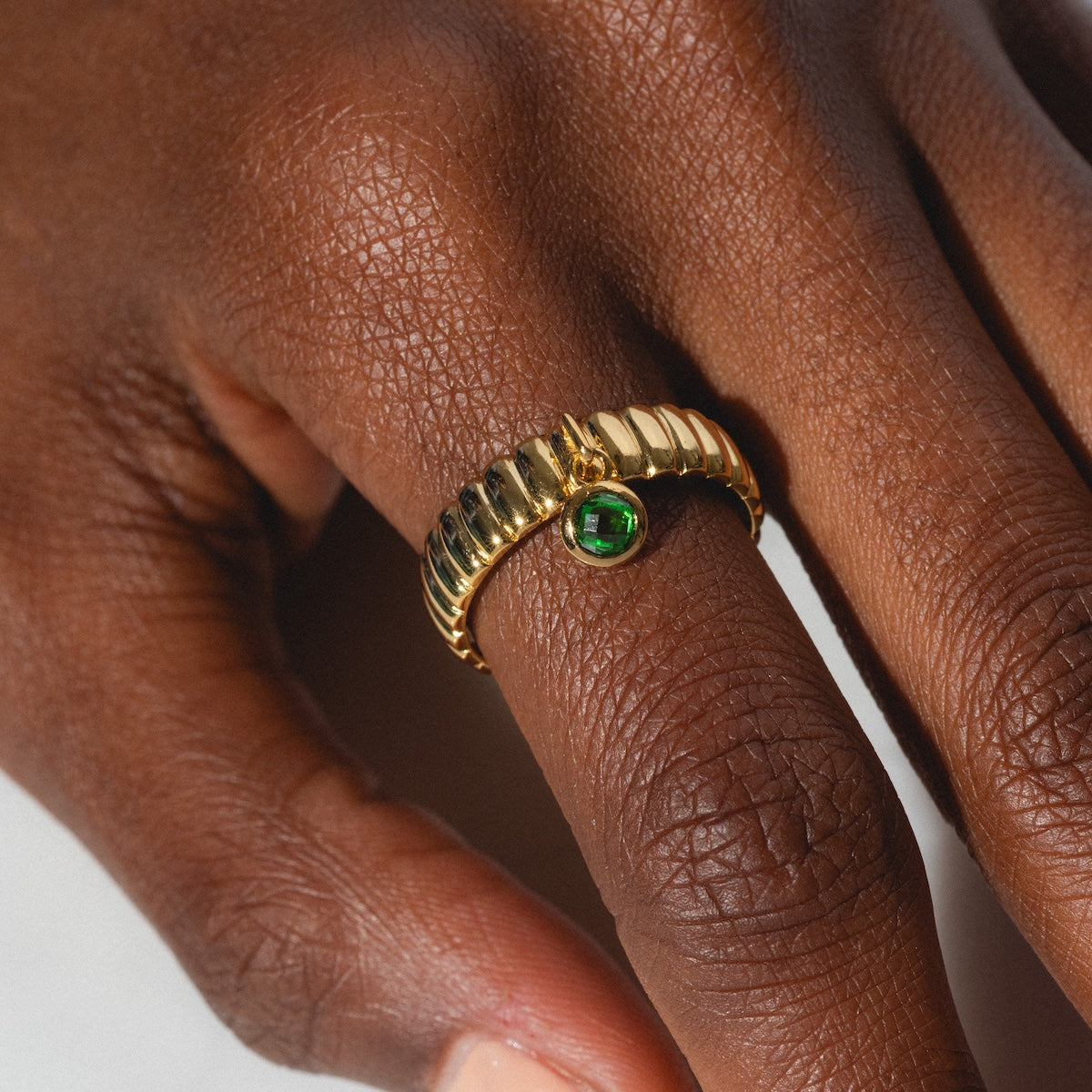 A close-up of a hand wearing the Bold Fluted Charm Ring, featuring a ridged 14k gold-plated band and a small round green gem in the center, shown on the middle finger.
