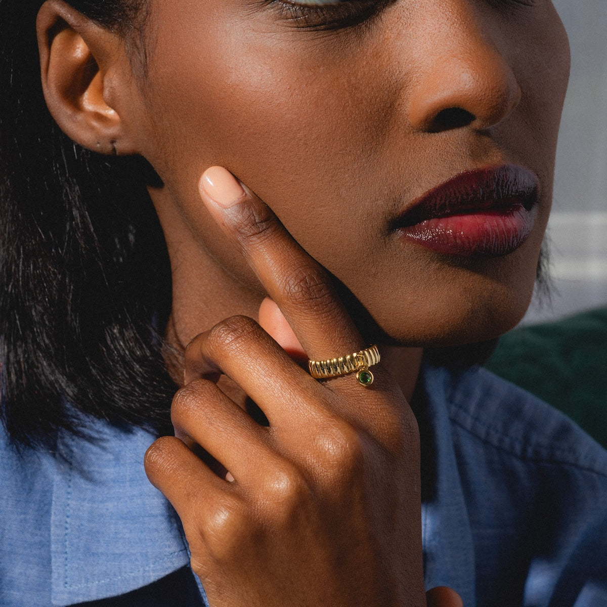 A woman with medium-dark skin wears the Bold Fluted Charm Ring, 14k gold plated with a green gemstone. She touches her cheek, highlighting her glossy red lipstick and blue collared shirt.